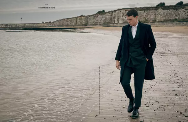 Man in dark suit and long coast walking along beach close to waters edge. Chalk cliffs seen in the background